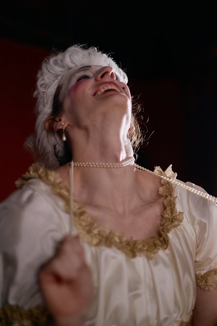About A woman dressed in a vintage white and gold dress with pearl jewelry is joyfully laughing, looking up.