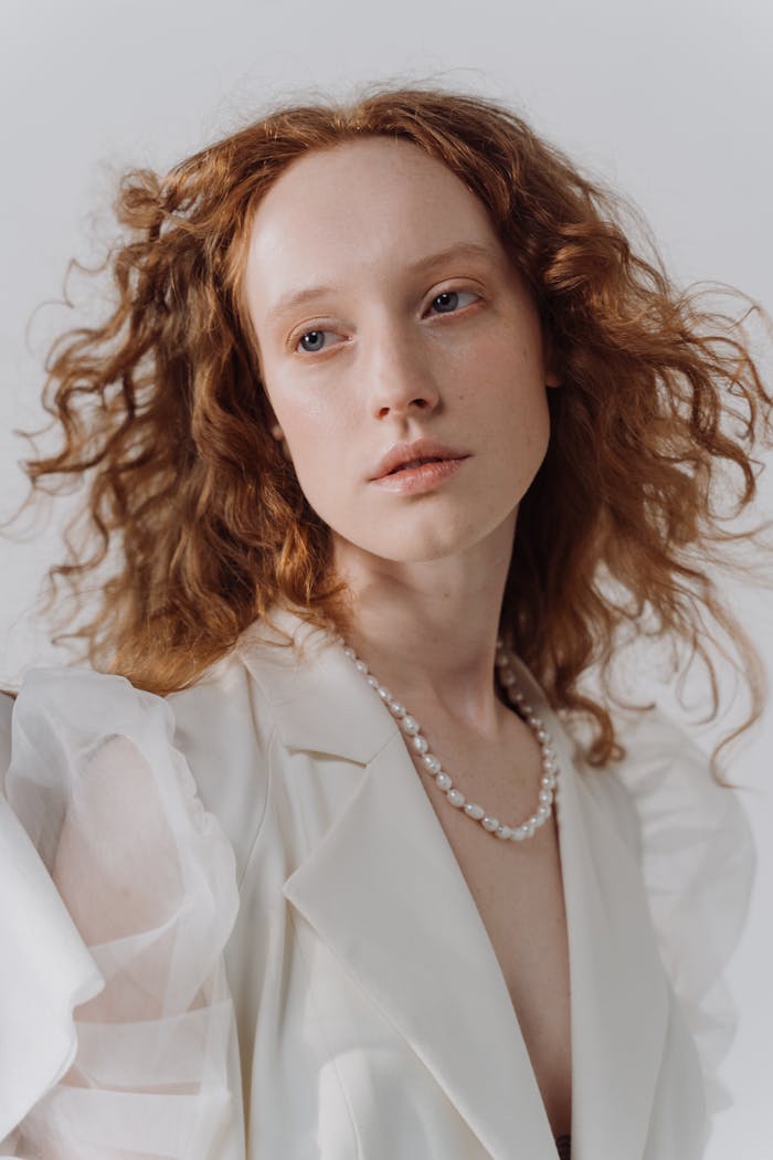 Home Portrait of a redhead woman with curly hair in a white blouse and pearl necklace on a white background.